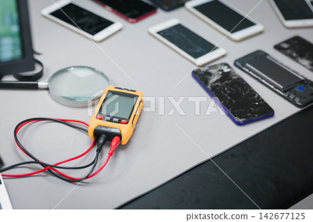 Technician workstation with damaged smart phones, a digital multimeter, test leads and magnifying glass for mobile phone repair and diagnostics. Technician workstation with damaged smart phones, a digital multimeter, test leads and magnifying glass for mobile phone repair and diagnostics. 142677125