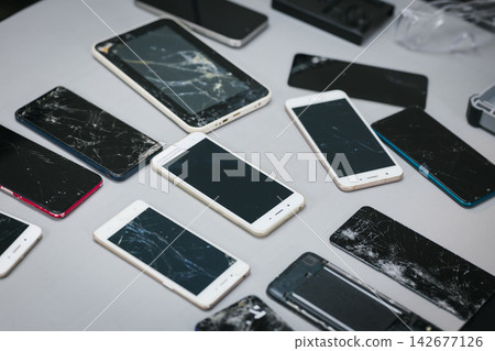 Collection of broken smart phones with cracked screens scattered on a table, device damage, repair needs, and electronic waste. Collection of broken smart phones with cracked screens scattered on a table, device damage, repair needs, and electronic waste. 142677126