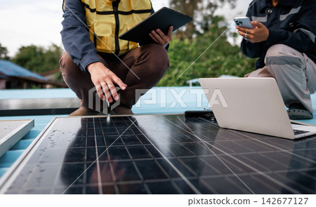 Two workers inspect solar panels on a roof, using a laptop, tablet and smartphone for renewable energy system monitoring. Two workers inspect solar panels on a roof, using a laptop, tablet and smartphone for renewable energy system monitoring. 142677127