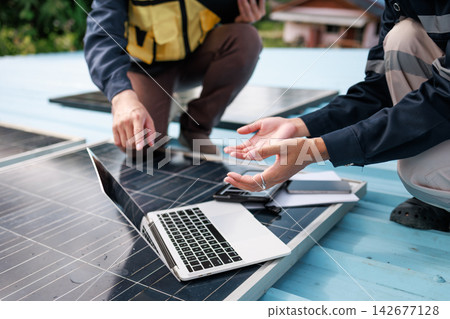 Two technicians inspect solar panels on a rooftop beside a laptop, planning renewable energy installation and maintenance outdoors. Two technicians inspect solar panels on a rooftop beside a laptop, planning renewable energy installation and maintenance outdoors. 142677128