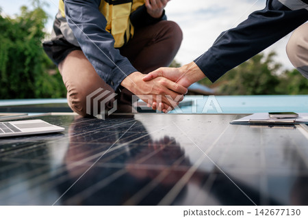 Two solar energy workers shake hands over rooftop photovoltaic panels, with a laptop and smartphone nearby, successful partnership and clean power project work. 142677130