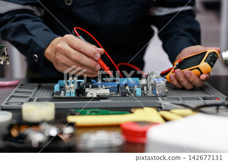 Close-up of a technician testing a computer circuit board with a digital multimeter during electronic device repair at a workbench. Close-up of a technician testing a computer circuit board with a digital multimeter during electronic device repair at a workbench. 142677131