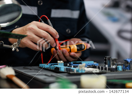 Close-up of a technician using a digital multimeter to test an electronic circuit board during computer repair in a workshop. 142677134