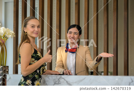 Happy female guest giving thumbs up at hotel reception front desk while receptionist provides assistance Happy female guest giving thumbs up at hotel reception front desk while receptionist provides assistance 142678319