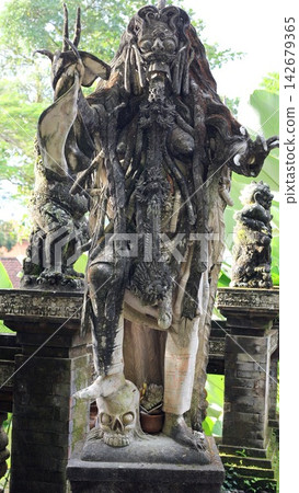 Stone demon statue stands on skull pedestal, menacing guardian with open mouth and elaborate carvings, aged Balinese style sculpture framed by green plants and shrine pillars, ornate carved details. Stone demon statue stands on skull pedestal, menacing guardian with open mouth and elaborate carvings, aged Balinese style sculpture framed by green plants and shrine pillars, ornate carved details. 142679365
