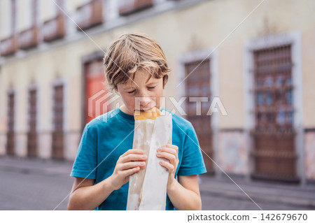 Boy eating a golden molote poblano traditional fried Mexican empanada outdoors on a street in Puebla Mexico with colonial city background paper wrapper local street food authentic travel snack. Boy eating a golden molote poblano traditional fried Mexican empanada outdoors on a street in Puebla Mexico with colonial city background paper wrapper local street food authentic travel snack. 142679670