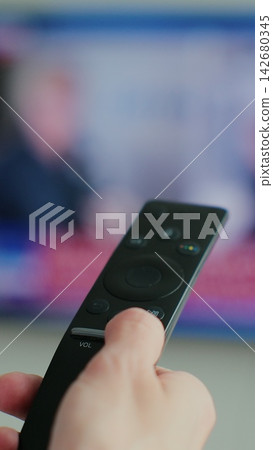 Vertical video. Woman hand switches TV remote control with finger, viewing of news, TV programs, modern technology for controlling equipment at a distance. Blurred TV in the background. Close up view. Vertical video. Woman hand switches TV remote control with finger, viewing of news, TV programs, modern technology for controlling equipment at a distance. Blurred TV in the background. Close up view. 142680345