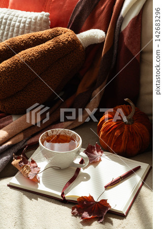 Cozy autumn scene with warm blanket, pumpkin decor, cup of tea and open notebook for reflection and journaling amidst fallen leaves and soft light 142683296