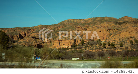 Orange badlands with ancient cave dwellings and sparse vegetation contrasting with a bright blue sky Orange badlands with ancient cave dwellings and sparse vegetation contrasting with a bright blue sky 142683423