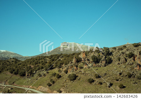 Mountain landscape showcasing rocky hills, scattered trees, and a distant snow-capped peak, providing a tranquil outdoor view Mountain landscape showcasing rocky hills, scattered trees, and a distant snow-capped peak, providing a tranquil outdoor view 142683426
