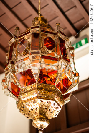 Ornate arabian lantern hanging, decorative islamic style chandelier with intricate amber stained glass and carved brass metalwork Ornate arabian lantern hanging, decorative islamic style chandelier with intricate amber stained glass and carved brass metalwork 142683567