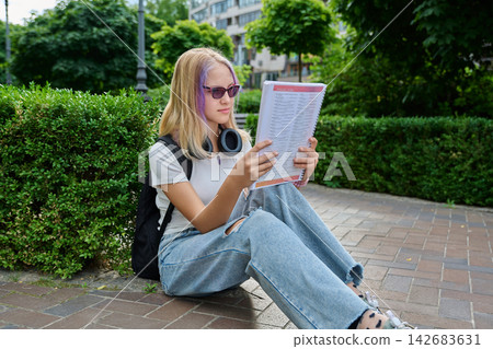 Teenage girl wearing sunglasses reading book sitting on pavement in park Teenage girl wearing sunglasses reading book sitting on pavement in park 142683631