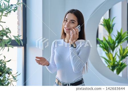 Smiling young woman talking on smartphone looking at camera in modern interior Smiling young woman talking on smartphone looking at camera in modern interior 142683644