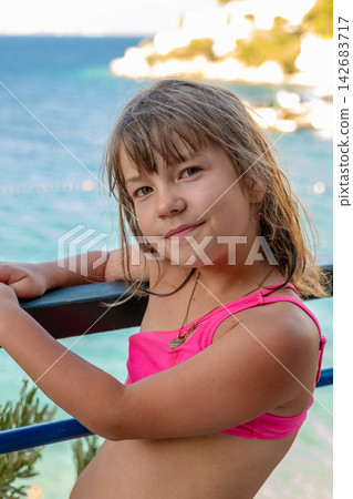 Smiling young girl in pink swimsuit posing by seaside railing Smiling young girl in pink swimsuit posing by seaside railing 142683717