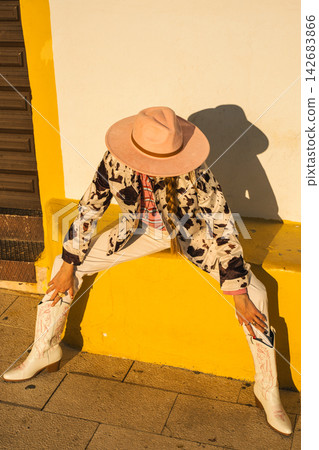 Stylish person in cowboy hat and boots sitting on yellow wall in sunlit urban setting Stylish person in cowboy hat and boots sitting on yellow wall in sunlit urban setting 142683866