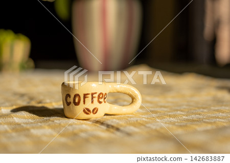 Tiny ceramic coffee mug on checkered tablecloth in sunlit kitchen decor Tiny ceramic coffee mug on checkered tablecloth in sunlit kitchen decor 142683887