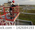 Industrial worker on elevated red platform handles maintenance gear, concentrated and steady in utility style setting. Worker wears red hard hat and safety harness above power lines and substation. 142684193