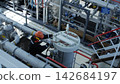 Technician repairs pipe valve in busy processing plant, skilled worker in protective gear leans over round unit, with steel framework, control lines, and heavy industrial equipment. 142684197