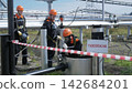 Three adult male technicians in orange helmets and protective suits examine gas piping at industrial facility, while one technician kneels at hatch. Hazard warning sign and barrier tape mark 142684201