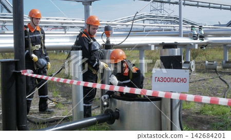 Three adult male technicians in orange helmets and protective suits examine gas piping at industrial facility, while one technician kneels at hatch. Hazard warning sign and barrier tape mark Three adult male technicians in orange helmets and protective suits examine gas piping at industrial facility, while one technician kneels at hatch. Hazard warning sign and barrier tape mark 142684201