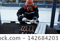 Maintenance worker kneels beside oversized wrench in industrial plant, wearing hard hat, eye protection, and gloves. Worker in protective gear handles tools on metal grating with intent focus. 142684203