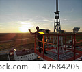 Facility maintenance worker stands on elevated platform at sunset, wearing helmet and workwear. He raises one hand beside mounted lights, cables, and tower frame above low buildings and open land. 142684205