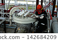 Industrial worker with concentrated expression adjusts bolted machinery in futuristic style plant, wearing helmet, goggles, and gloves. Worker uses wrench beside large valve, pipes, and wrapped parts. 142684209