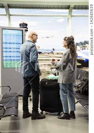 Smiling older adults, observing flight board, with departures timetable and route check information in airport terminal. Mature couple reviewing flight update while enjoying freedom. Smiling older adults, observing flight board, with departures timetable and route check information in airport terminal. Mature couple reviewing flight update while enjoying freedom. 142685639