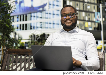 Black adult sitting on a bench scrolling through web page on laptop, casual urban lifestyle contrasted with the modern downtown buildings and dynamic pedestrian scenery behind him. Black adult sitting on a bench scrolling through web page on laptop, casual urban lifestyle contrasted with the modern downtown buildings and dynamic pedestrian scenery behind him. 142685661