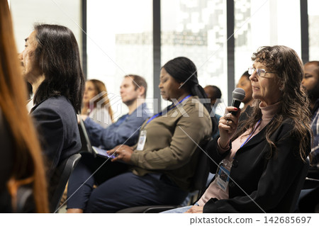 Senior woman in audience using microphone to ask multiple questions and engage in business panel discussion, pitch during international conference summit. Development and training. Senior woman in audience using microphone to ask multiple questions and engage in business panel discussion, pitch during international conference summit. Development and training. 142685697