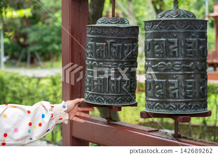 Cropped shot view of woman hand try to spinning a prayer wheel in Tibetan Buddhist monastery. Prayer wheels are devices for spreading spiritual blessings and well being. Cropped shot view of woman hand try to spinning a prayer wheel in Tibetan Buddhist monastery. Prayer wheels are devices for spreading spiritual blessings and well being. 142689262