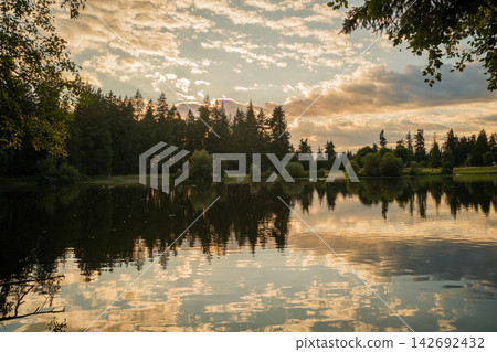 Scenic view of a calm lake reflecting a pine forest and golden sunset sky with clouds Scenic view of a calm lake reflecting a pine forest and golden sunset sky with clouds 142692432