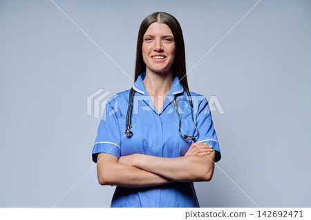 Confident female nurse in blue uniform with stethoscope standing with arms crossed against grey background Confident female nurse in blue uniform with stethoscope standing with arms crossed against grey background 142692471