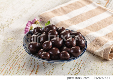 Glossy dark chocolate coated candies in blue and white ceramic bowl on rustic wooden surface with striped cloth and purple flower Glossy dark chocolate coated candies in blue and white ceramic bowl on rustic wooden surface with striped cloth and purple flower 142692647