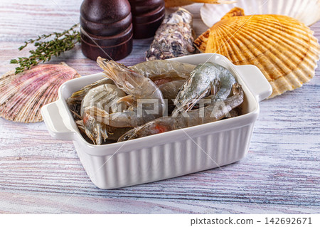 Raw fresh shrimp in white ceramic dish, green thyme sprig, variety of seashells, wooden grinders on rustic light wood background, seafood preparation Raw fresh shrimp in white ceramic dish, green thyme sprig, variety of seashells, wooden grinders on rustic light wood background, seafood preparation 142692671