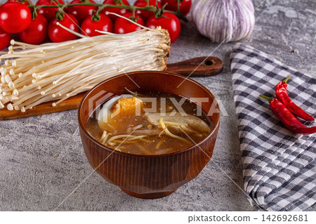 Bowl of miso ramen with soft boiled egg, enoki mushrooms and chicken in rich broth, surrounded by cherry tomatoes, garlic, red chili peppers and checkered napkin on gray stone surface Bowl of miso ramen with soft boiled egg, enoki mushrooms and chicken in rich broth, surrounded by cherry tomatoes, garlic, red chili peppers and checkered napkin on gray stone surface 142692681