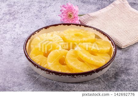 Sliced pineapple rings in ceramic bowl on textured gray surface with pink flower and woven napkin background Sliced pineapple rings in ceramic bowl on textured gray surface with pink flower and woven napkin background 142692776