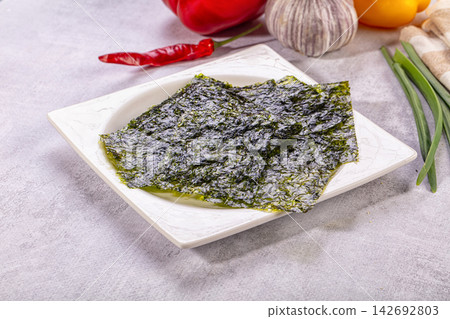 Dark green nori seaweed sheets on white ceramic plate with red pepper, garlic, and green onions in background Dark green nori seaweed sheets on white ceramic plate with red pepper, garlic, and green onions in background 142692803