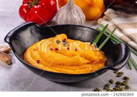Creamy orange pumpkin soup in black bowl garnished with pumpkin seeds and red peppercorns, surrounded by fresh vegetables and garlic Creamy orange pumpkin soup in black bowl garnished with pumpkin seeds and red peppercorns, surrounded by fresh vegetables and garlic 142692804