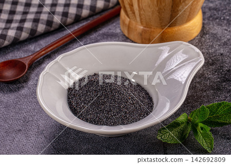Poppy seeds in white ceramic bowl with fresh mint leaves, wooden spoon and checkered cloth on dark textured surface Poppy seeds in white ceramic bowl with fresh mint leaves, wooden spoon and checkered cloth on dark textured surface 142692809