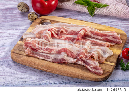 Raw bacon strips on wooden cutting board with quail eggs, tomato, and basil on rustic painted surface Raw bacon strips on wooden cutting board with quail eggs, tomato, and basil on rustic painted surface 142692843
