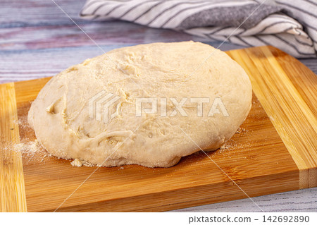 Homemade yeast dough for bread or pizza resting on rustic bamboo cutting board, light flour coating, vibrant blue and purple background 142692890