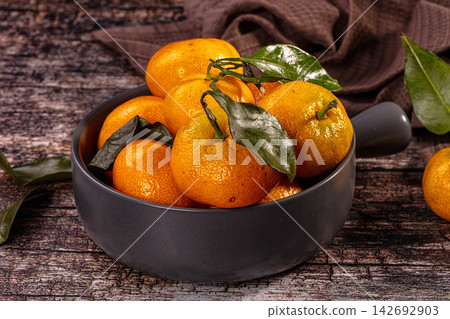 Fresh tangerines with green leaves in matte black bowl on rustic wooden surface, warm orange tones and natural texture Fresh tangerines with green leaves in matte black bowl on rustic wooden surface, warm orange tones and natural texture 142692903