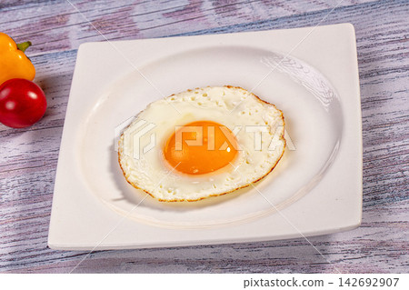 Fried egg with runny yolk on white square plate, beside yellow pepper and red tomato on rustic wood surface Fried egg with runny yolk on white square plate, beside yellow pepper and red tomato on rustic wood surface 142692907
