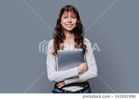 Smiling young female student holding laptop looking at camera on grey background Smiling young female student holding laptop looking at camera on grey background 142692996