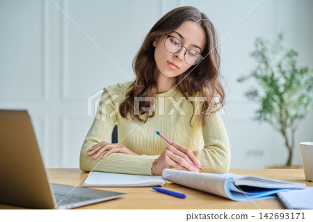Focused female student taking notes at desk with laptop front view Focused female student taking notes at desk with laptop front view 142693171