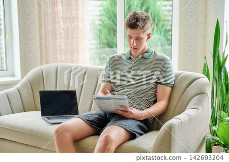 Focused 18-year-old student writing in notebook sitting on sofa with laptop Focused 18-year-old student writing in notebook sitting on sofa with laptop 142693204