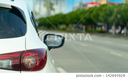 Rear view of a white car driving on a city street under bright daytime conditions. Plant ornamental trees along the entire length of the road. Blurred of town for background under blue sky. Rear view of a white car driving on a city street under bright daytime conditions. Plant ornamental trees along the entire length of the road. Blurred of town for background under blue sky. 142693929