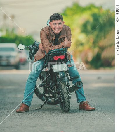 Smiling latin motorcyclist leaning against his motorcycle and looking at the camera Smiling latin motorcyclist leaning against his motorcycle and looking at the camera 142693991