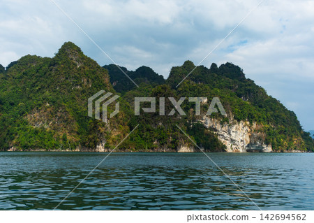 Rocky mountains, lush with trees, rise from the azure water, their cliffs catching the afternoon sun, set against a sky filled with clouds. Rocky mountains, lush with trees, rise from the azure water, their cliffs catching the afternoon sun, set against a sky filled with clouds. 142694562